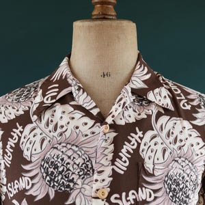 Peut inclure: Une chemise &agrave; manches courtes marron avec un imprim&eacute; floral rose et blanc. L'imprim&eacute; repr&eacute;sente des ananas, des feuilles et le texte "Hawaii Island".
