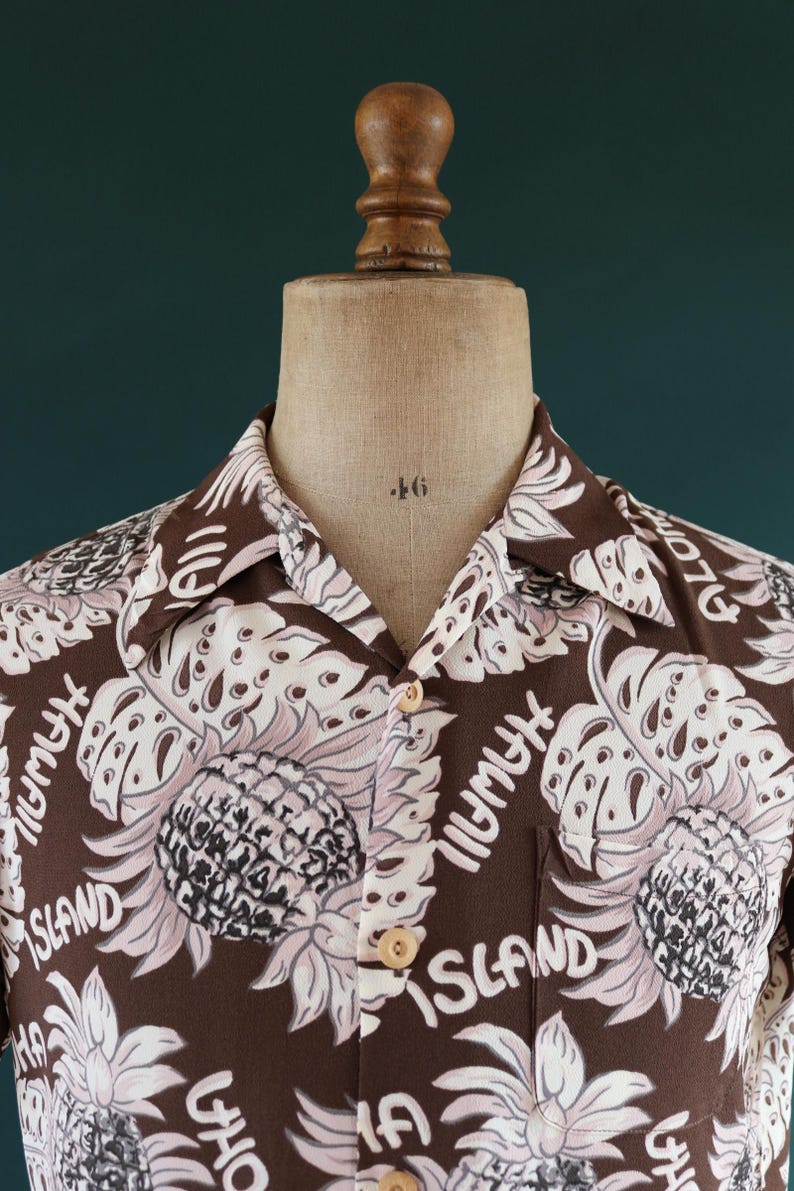 Peut inclure: Une chemise &agrave; manches courtes marron avec un imprim&eacute; floral rose et blanc. L'imprim&eacute; repr&eacute;sente des ananas, des feuilles et le texte "Hawaii Island".