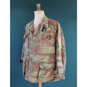 May include: A camouflage jacket in brown, green, and beige tones, shown on a mannequin. The jacket has several pockets, a button-up design, and long sleeves. The fabric displays a unique pattern, often seen in military-style clothing.