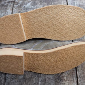 May include: Two olive green suede shoes with light brown soles are displayed on a wooden surface. The soles have a textured, crepe-like appearance. The stitching is visible along the sides of the shoes.
