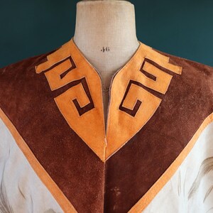 May include: A white suede poncho with brown suede trim and a geometric pattern around the neckline. The poncho has a V-neck and is decorated with a brown and white bird design.