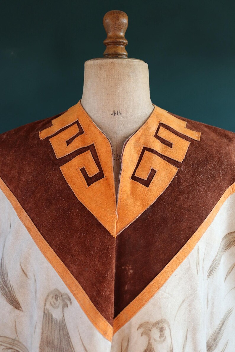 May include: A white suede poncho with brown suede trim and a geometric pattern around the neckline. The poncho has a V-neck and is decorated with a brown and white bird design.