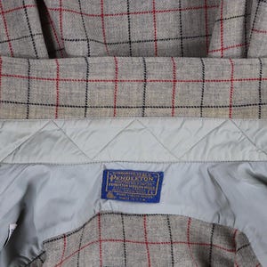 May include: Close-up of a gray plaid jacket interior with red and black grid lines. The jacket has a light blue quilted lining and a blue label with white text. The label reads "Pendleton".