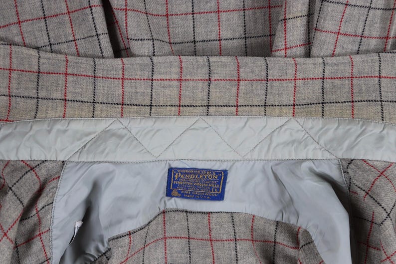 May include: Close-up of a gray plaid jacket interior with red and black grid lines. The jacket has a light blue quilted lining and a blue label with white text. The label reads "Pendleton".