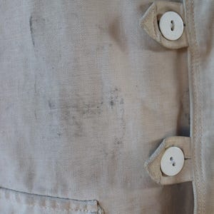 May include: Close-up of a beige fabric garment with visible staining. The garment features two white buttons and a pocket. The fabric appears to be a sturdy canvas or similar material, with a visible weave.