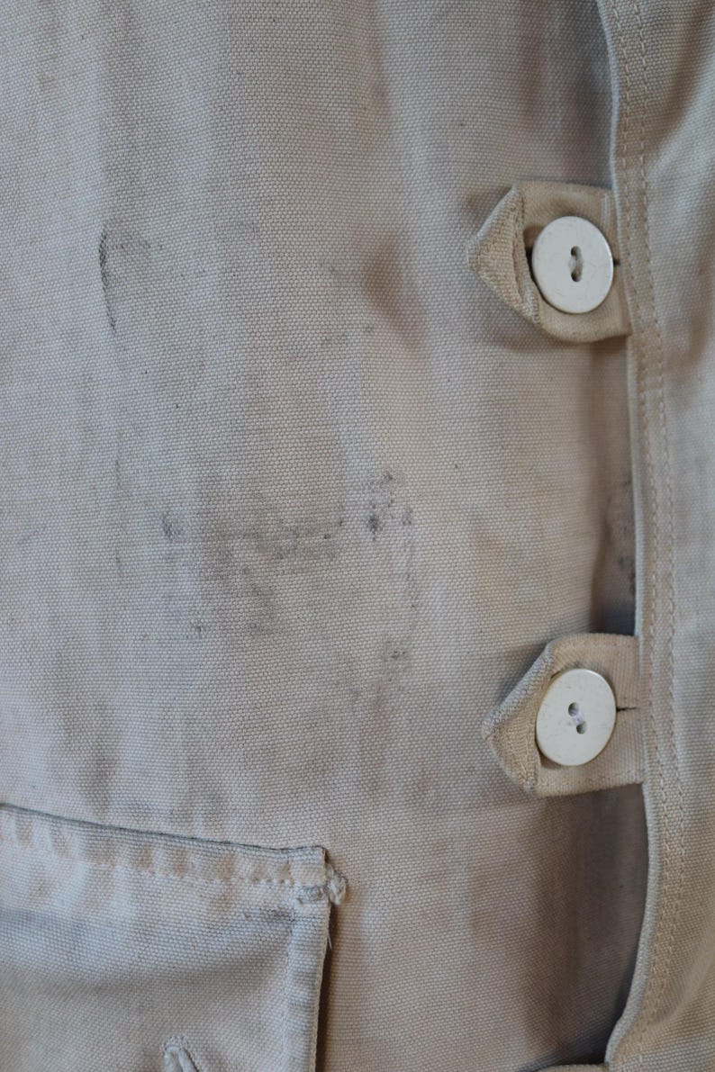 May include: Close-up of a beige fabric garment with visible staining. The garment features two white buttons and a pocket. The fabric appears to be a sturdy canvas or similar material, with a visible weave.