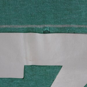 May include: Close-up of a teal fabric with a white, paper-like shape cut out in the shape of the number seven. The fabric has a subtle texture and a horizontal white line.