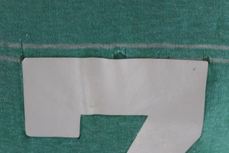 May include: Close-up of a teal fabric with a white, paper-like shape cut out in the shape of the number seven. The fabric has a subtle texture and a horizontal white line.