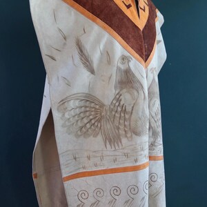 May include: A white leather poncho with brown suede trim and a rooster design. The poncho has a fringe border and a geometric pattern around the neck.