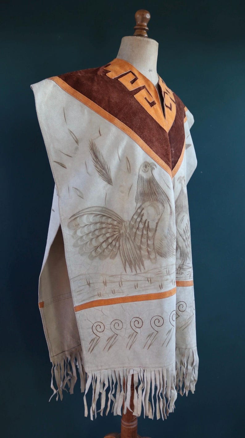 May include: A white leather poncho with brown suede trim and a rooster design. The poncho has a fringe border and a geometric pattern around the neck.
