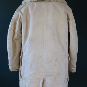 May include: Cream-colored winter coat with a shearling collar. The coat has a long length, two pockets, and a split at the back hem. The collar is a light beige color. The coat is made of a thick, durable material.