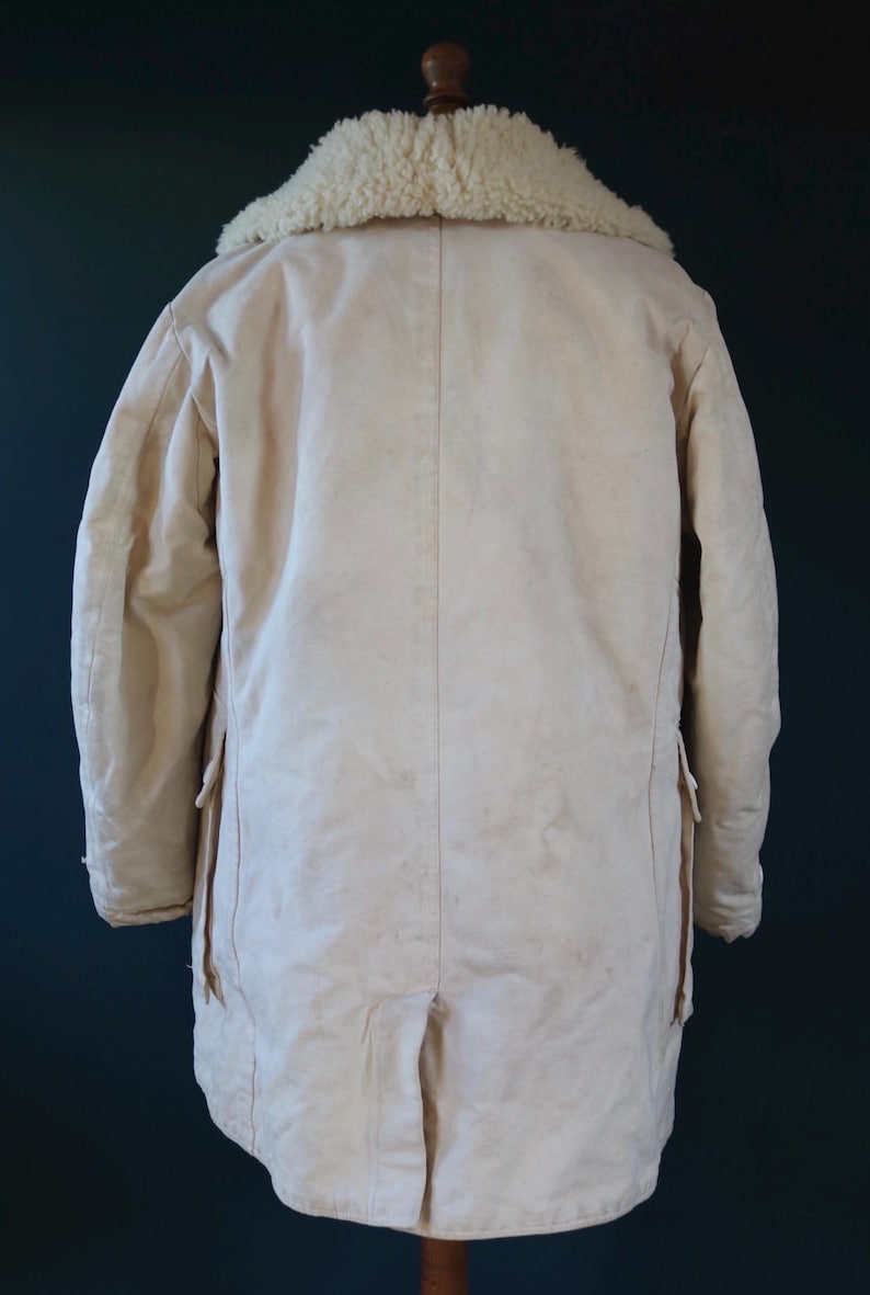 May include: Cream-colored winter coat with a shearling collar. The coat has a long length, two pockets, and a split at the back hem. The collar is a light beige color. The coat is made of a thick, durable material.