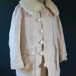 May include: Cream-colored vintage coat with a fluffy, off-white shearling collar. The coat features large white buttons, two front pockets, and buttoned cuffs. The coat is displayed on a wooden mannequin.