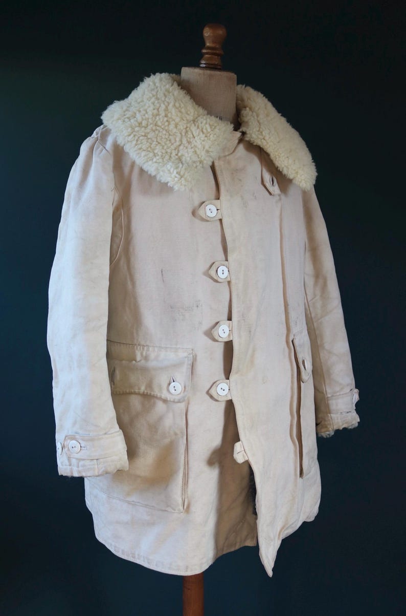 May include: Cream-colored vintage coat with a fluffy, off-white shearling collar. The coat features large white buttons, two front pockets, and buttoned cuffs. The coat is displayed on a wooden mannequin.