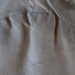 May include: Close-up of a beige canvas fabric with a visible seam and a section of fluffy, off-white material. The canvas has a textured appearance, with a small blue mark. The item appears to be part of a garment.