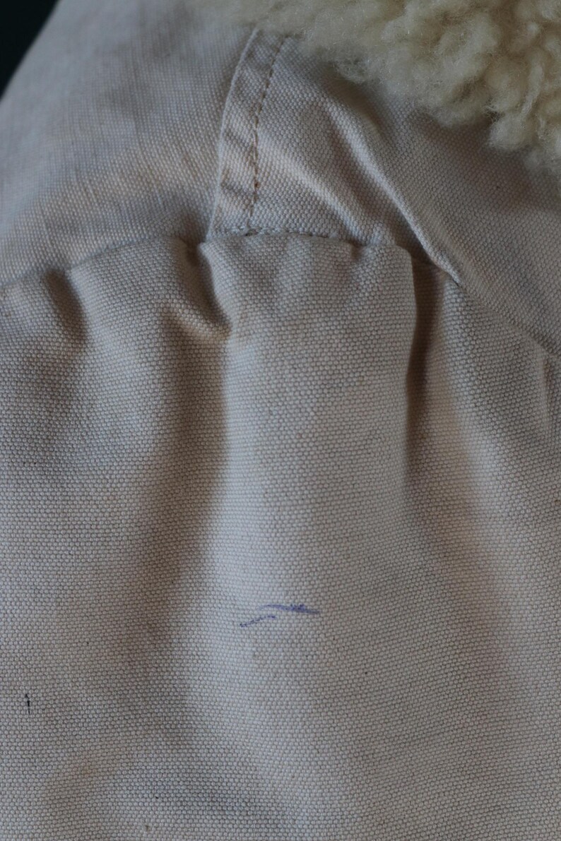 May include: Close-up of a beige canvas fabric with a visible seam and a section of fluffy, off-white material. The canvas has a textured appearance, with a small blue mark. The item appears to be part of a garment.