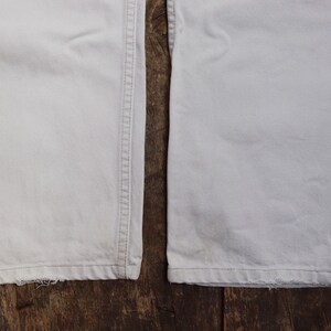 May include: Close-up of the bottom hems of two pairs of white pants. The pants have a straight leg and are made of a lightweight fabric.