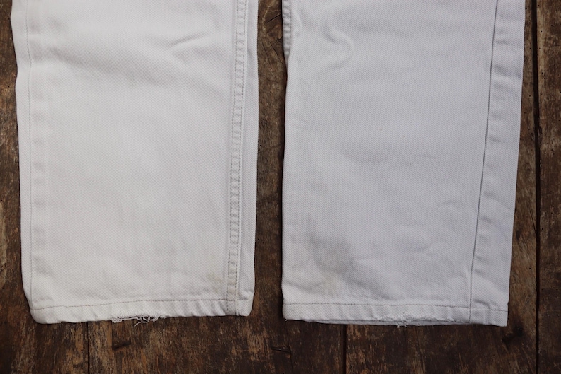 May include: Close-up of the bottom hems of two pairs of white pants. The pants have a straight leg and are made of a lightweight fabric.