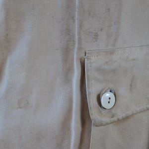 May include: Close-up of tan fabric with a small pocket and a white button. The fabric has some stains and a textured appearance. The pocket is rectangular and stitched onto the fabric.