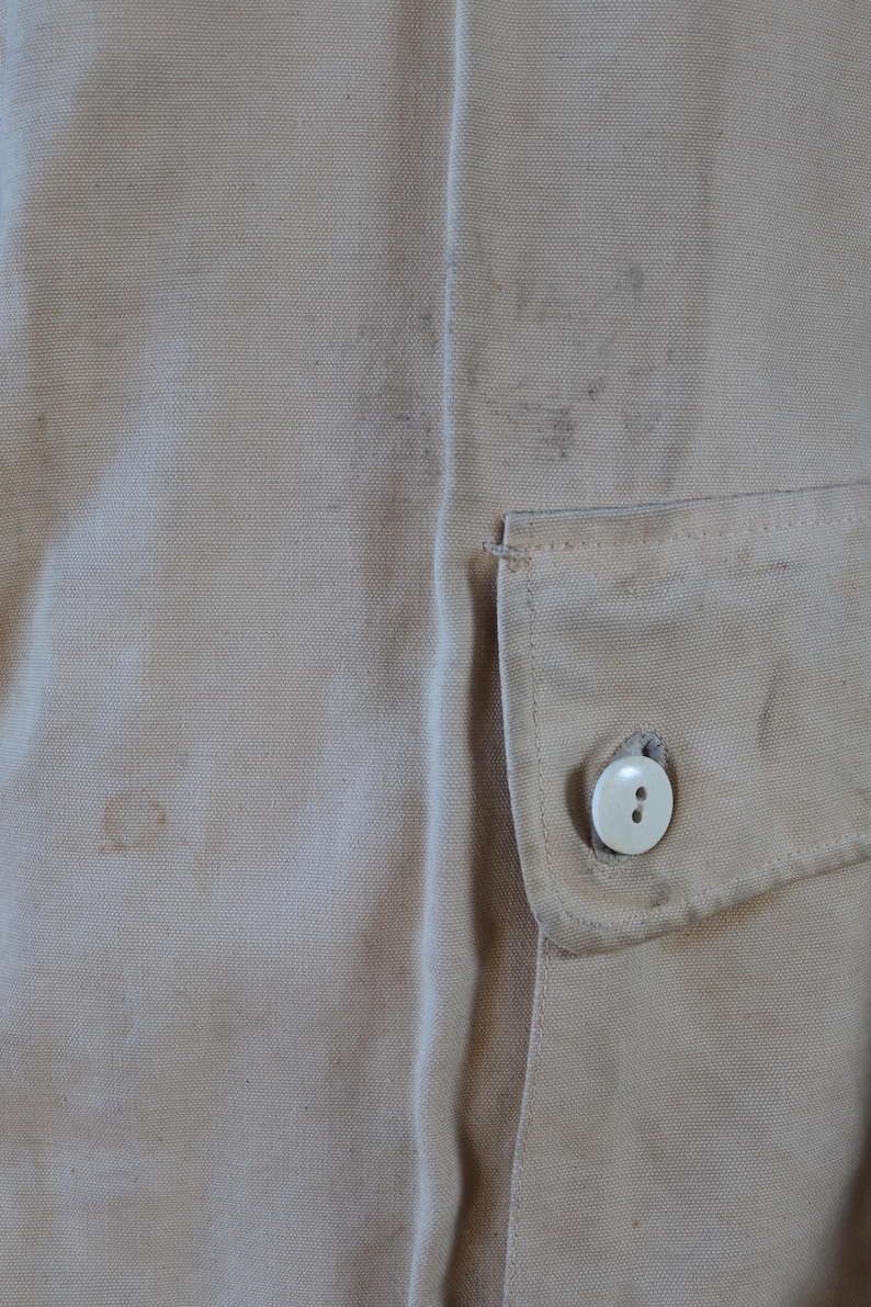 May include: Close-up of tan fabric with a small pocket and a white button. The fabric has some stains and a textured appearance. The pocket is rectangular and stitched onto the fabric.