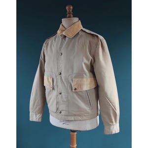 May include: A tan jacket with a corduroy collar and two front pockets. The jacket is buttoned up and displayed on a mannequin. The jacket has a classic, vintage style.