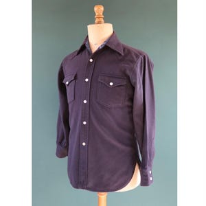 May include: A dark navy blue long-sleeved shirt with two chest pockets and white buttons. The shirt is displayed on a mannequin. The collar has a patterned lining. The cuffs also have white buttons.