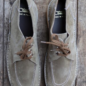 May include: A pair of olive green suede shoes with brown laces. The shoes have white stitching and are labeled "Made in Poland" inside. The shoes are displayed on a wooden surface.