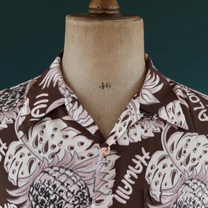 Peut inclure: Une chemise boutonn&eacute;e marron avec un motif floral blanc. La chemise a un col et des manches courtes. Le motif comprend de grandes feuilles et le mot "Plymouth" r&eacute;p&eacute;t&eacute; partout.