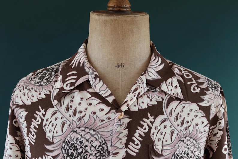 Peut inclure: Une chemise boutonn&eacute;e marron avec un motif floral blanc. La chemise a un col et des manches courtes. Le motif comprend de grandes feuilles et le mot "Plymouth" r&eacute;p&eacute;t&eacute; partout.