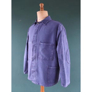 May include: A blue work jacket with a button closure, a collar, and three pockets. The jacket has an elbow patch. The jacket is displayed on a wooden mannequin.