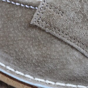 May include: Close-up of a gray suede shoe. The shoe features white stitching along the seams and a light blue accent near the sole. The texture of the suede is visible, showing a slightly worn appearance.