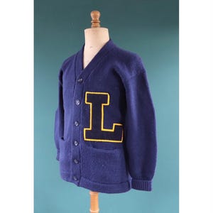 May include: A navy blue cardigan with a large yellow and black letter "L" patch. The sweater features a V-neck, button closure, and two front pockets. It is displayed on a mannequin. The cardigan is made of a knitted fabric.