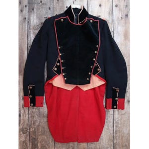 May include: A vintage military jacket in navy blue with a black velvet front panel and red detailing. It has gold buttons and a red tailcoat. The jacket is displayed against a wooden background.