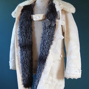 May include: Cream-colored vintage coat with a sheepskin collar and dark gray fur lining. The coat features button closures, pockets, and a strap across the chest. The coat is displayed on a mannequin.