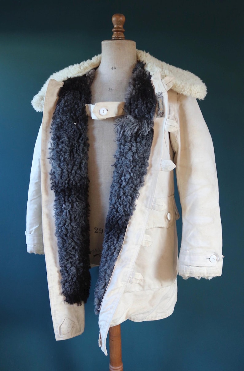 May include: Cream-colored vintage coat with a sheepskin collar and dark gray fur lining. The coat features button closures, pockets, and a strap across the chest. The coat is displayed on a mannequin.