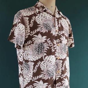 Peut inclure: Une chemise &agrave; manches courtes marron avec un motif de feuilles et d'ananas blancs. La chemise porte les mots "Aloha", "Hawaii" et "Island" imprim&eacute;s dessus.