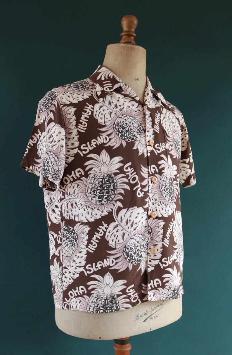 Peut inclure: Une chemise &agrave; manches courtes marron avec un motif de feuilles et d'ananas blancs. La chemise porte les mots "Aloha", "Hawaii" et "Island" imprim&eacute;s dessus.