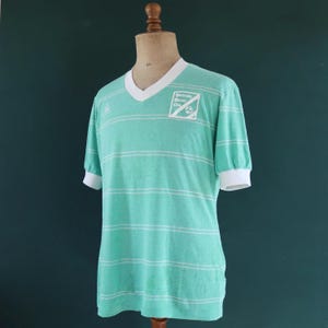 May include: A vintage teal and white striped soccer jersey on a mannequin. The jersey has a V-neck, white cuffs, and a logo that reads "Bethesda Soccer Club". The shirt is a light, breathable material.