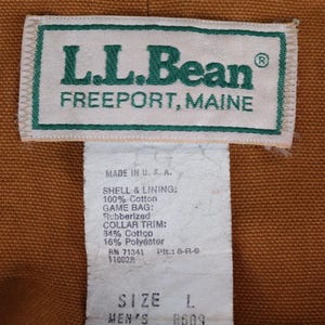 May include: Close-up of an L.L.Bean label, with text reading "FREEPORT, MAINE". The label is white with green text and trim. Below the label is a tag with product information, including size L and the color brown.