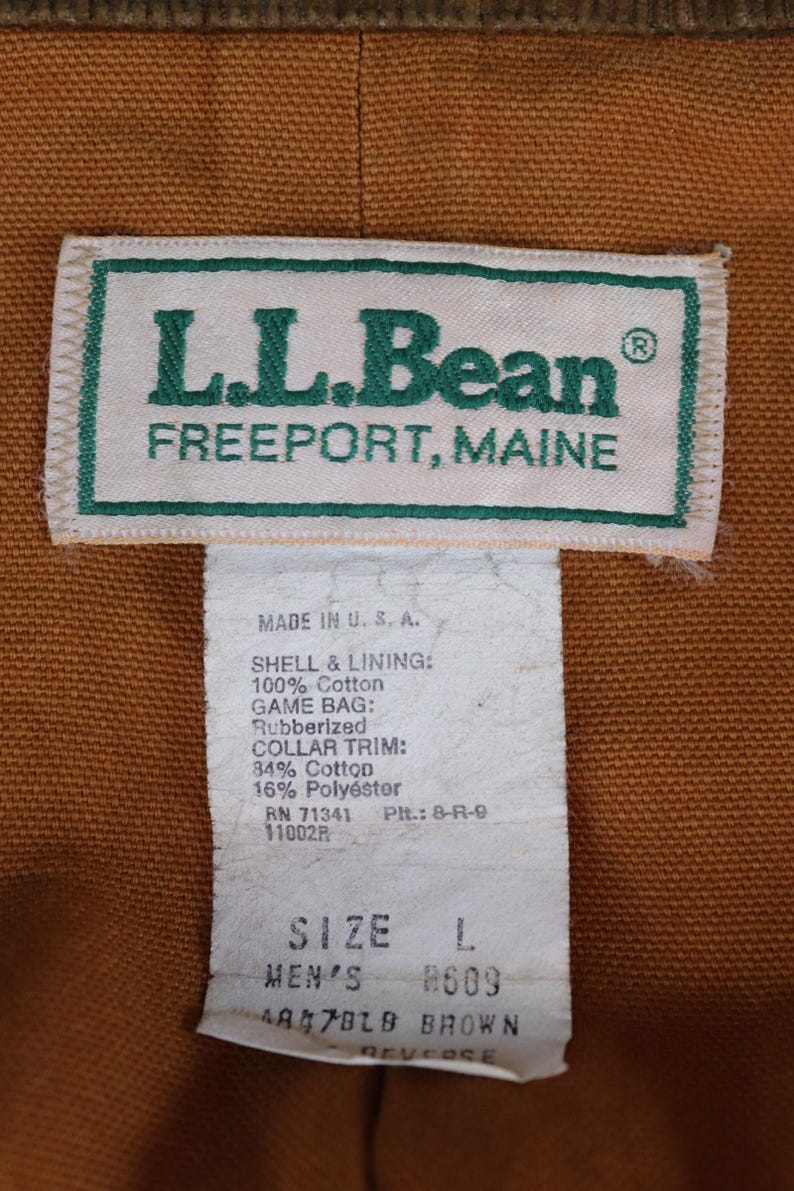 May include: Close-up of an L.L.Bean label, with text reading "FREEPORT, MAINE". The label is white with green text and trim. Below the label is a tag with product information, including size L and the color brown.