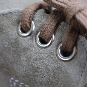 May include: Close-up of a light green suede shoe. The image highlights the shoe's brown laces threaded through silver eyelets. White stitching is visible along the edge of the shoe, adding a contrasting detail to the design.