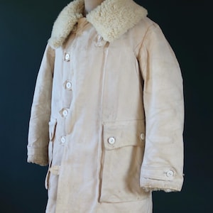 May include: Cream-colored vintage winter coat with a sheepskin collar. The coat features large white buttons, two front pockets, and a buttoned cuff. The coat is displayed on a wooden mannequin.
