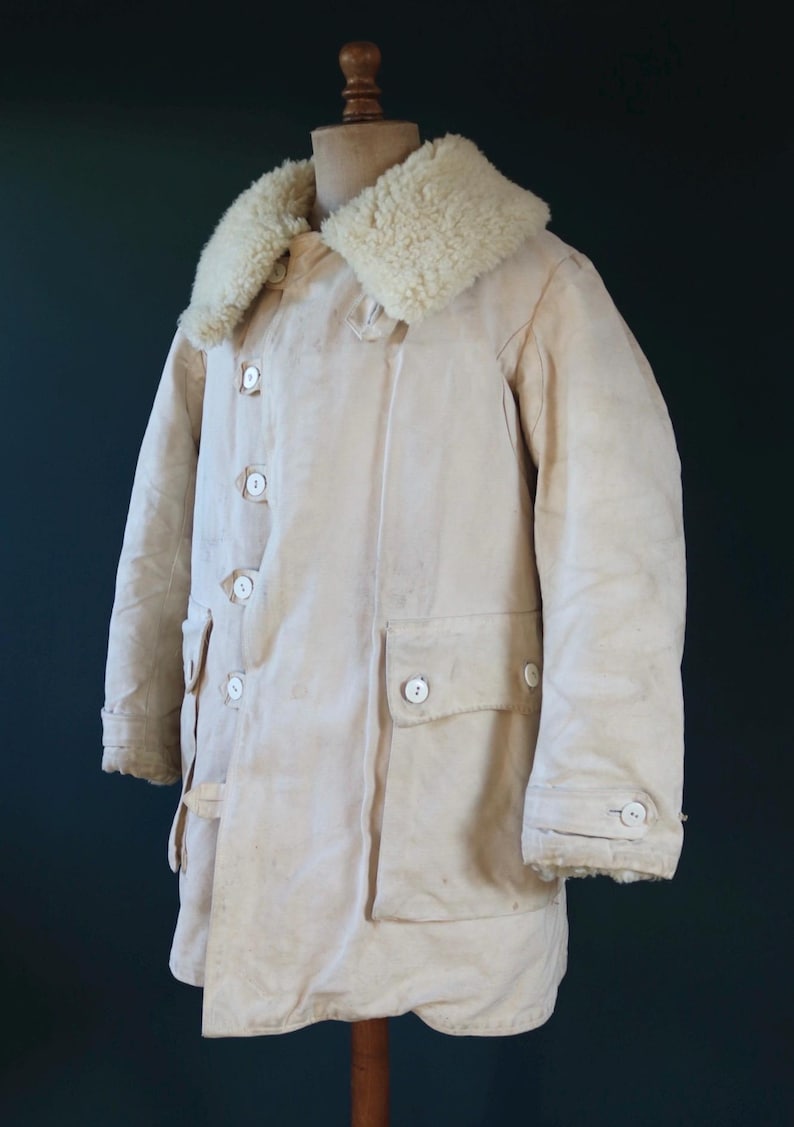 May include: Cream-colored vintage winter coat with a sheepskin collar. The coat features large white buttons, two front pockets, and a buttoned cuff. The coat is displayed on a wooden mannequin.