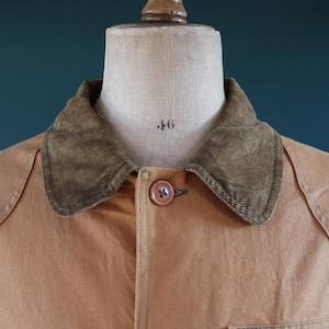 May include: Close-up of a tan work jacket on a mannequin. The jacket has a corduroy collar, a button closure, and a small zippered pocket. The mannequin is marked with the number 46.