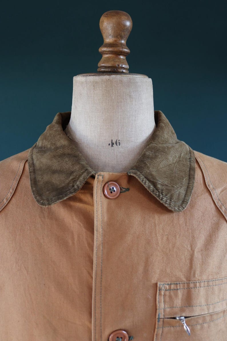 May include: Close-up of a tan work jacket on a mannequin. The jacket has a corduroy collar, a button closure, and a small zippered pocket. The mannequin is marked with the number 46.
