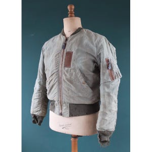 May include: A vintage olive green bomber jacket with a brown leather patch and zipper details. The jacket is displayed on a mannequin, showcasing its ribbed cuffs and waistband. The jacket has a classic, worn aesthetic.