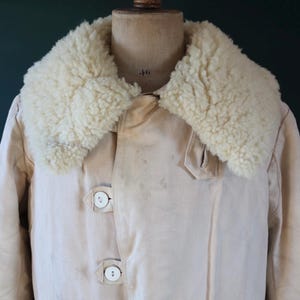 May include: Cream-colored coat with a fluffy, off-white shearling collar. The coat features three large, round, white buttons and a classic design. The garment is displayed on a vintage mannequin.