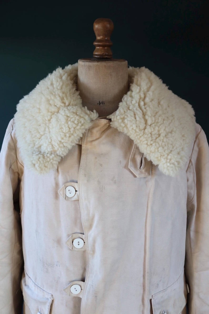 May include: Cream-colored coat with a fluffy, off-white shearling collar. The coat features three large, round, white buttons and a classic design. The garment is displayed on a vintage mannequin.