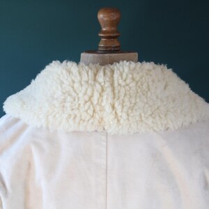 May include: Cream-colored jacket with a fluffy, off-white shearling collar. The jacket is displayed on a wooden mannequin head against a teal background. The collar is the focal point, offering a textural contrast to the smooth fabric.