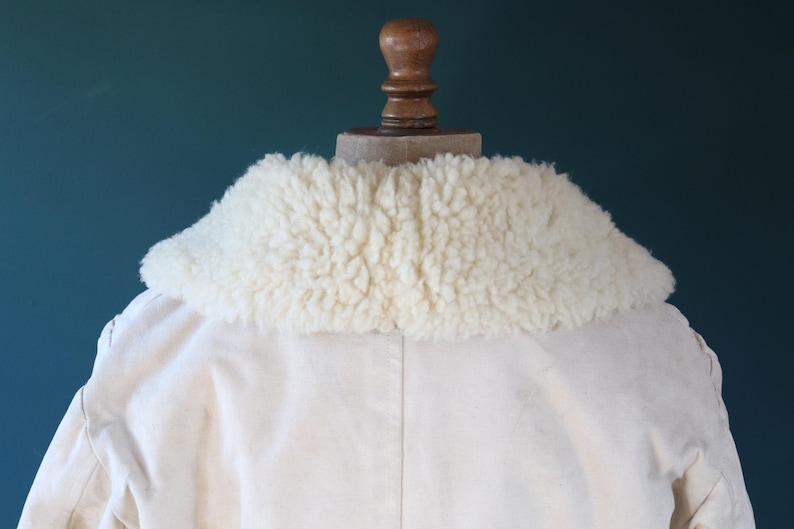 May include: Cream-colored jacket with a fluffy, off-white shearling collar. The jacket is displayed on a wooden mannequin head against a teal background. The collar is the focal point, offering a textural contrast to the smooth fabric.